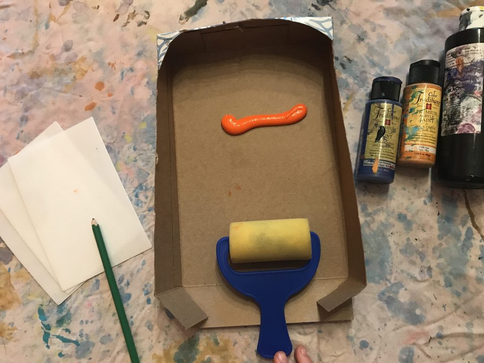 Arrangement of printmaking materials. At the center, a cardboard tray holds orange paint and a brayer. To either side, a pencil, paper, and paint tubes.