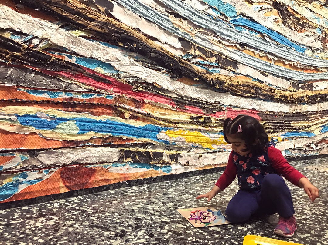 A child examines a piece of artwork on the floor, with Mark Bradford's Pickett's Charge in the background.