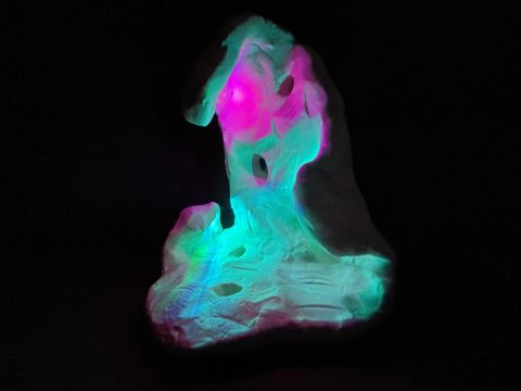 A white sculpture sits on a pitch black field, while blue pink and green light dance on it's surface.