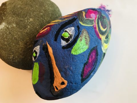 A long rock with a colorfully painted face and decorations along the rest of it's body leans against a smaller rock.