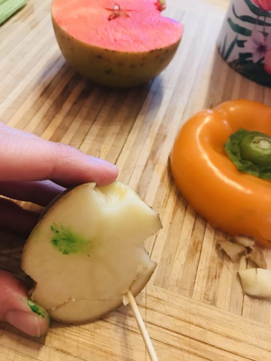 An arrangement of cut apple, orange pepper, and potato on a wood cutting board. A toothpick carves out the edges of the potato.