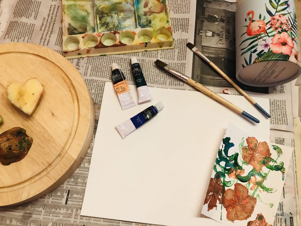 Tools and materials are staged for artmaking. A white paper rests on newspaper with tubes of paint and paintbrushes nearby. To the left, a wood board holds a potato cut in the shape of a heart.