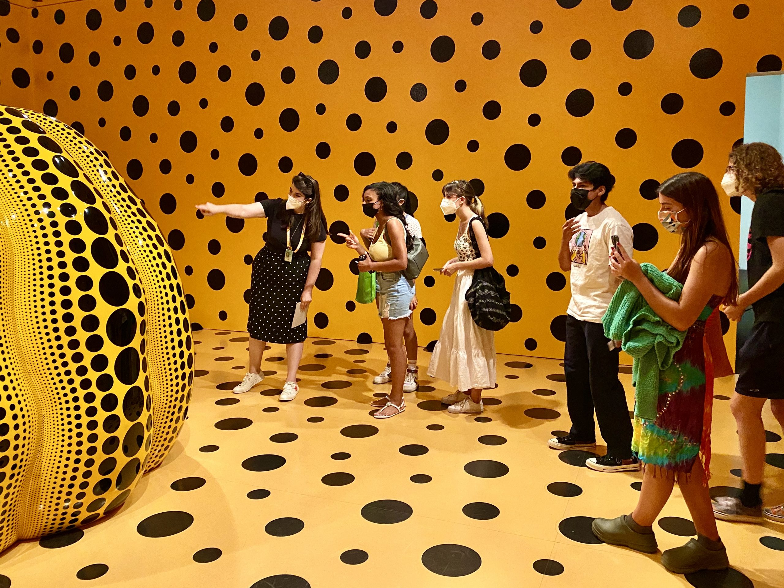 Teens in yellow painted room with black polka dots looking at a large yellow and black polka dotted pumpkin sculpture