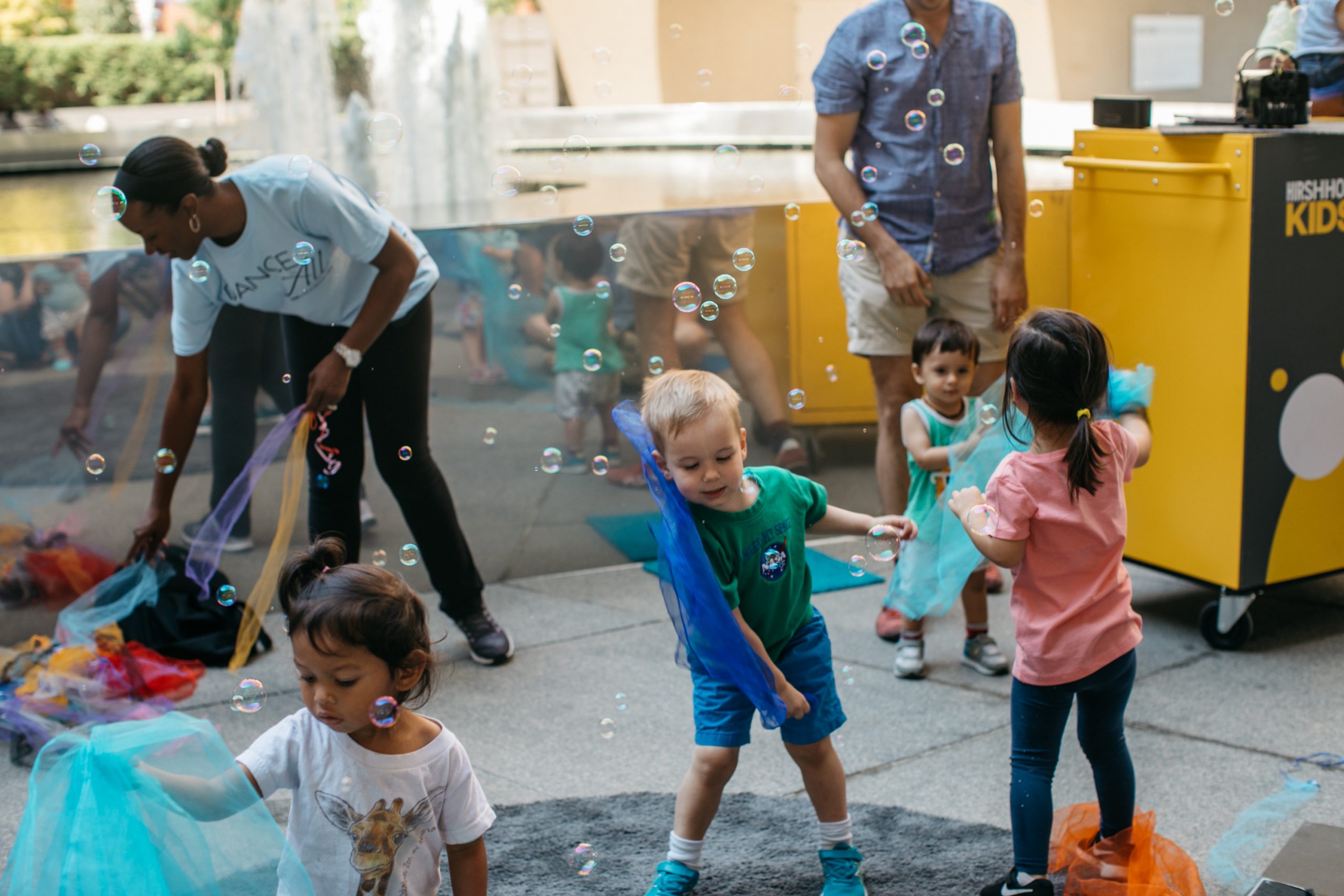 A group of children and adults play in the Hirshhorn plaza, the fountain behind them.