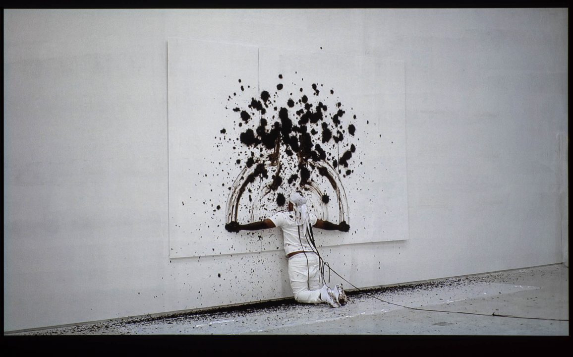 Artist dressed in white painting with body on white canvas hung in a white studio