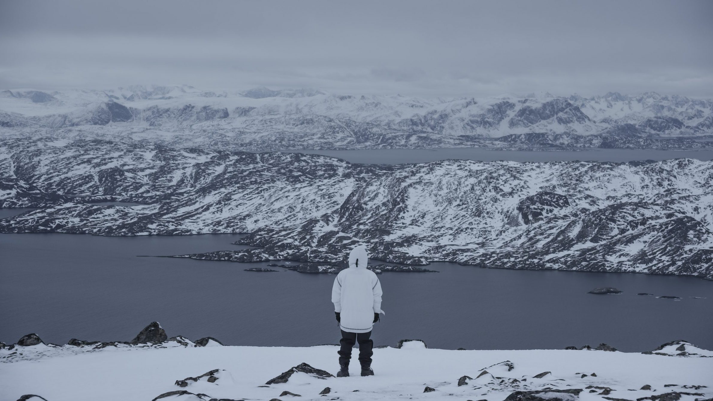 Snowy landscape of mountains and a body of water with a person standing on a ridge overlooking