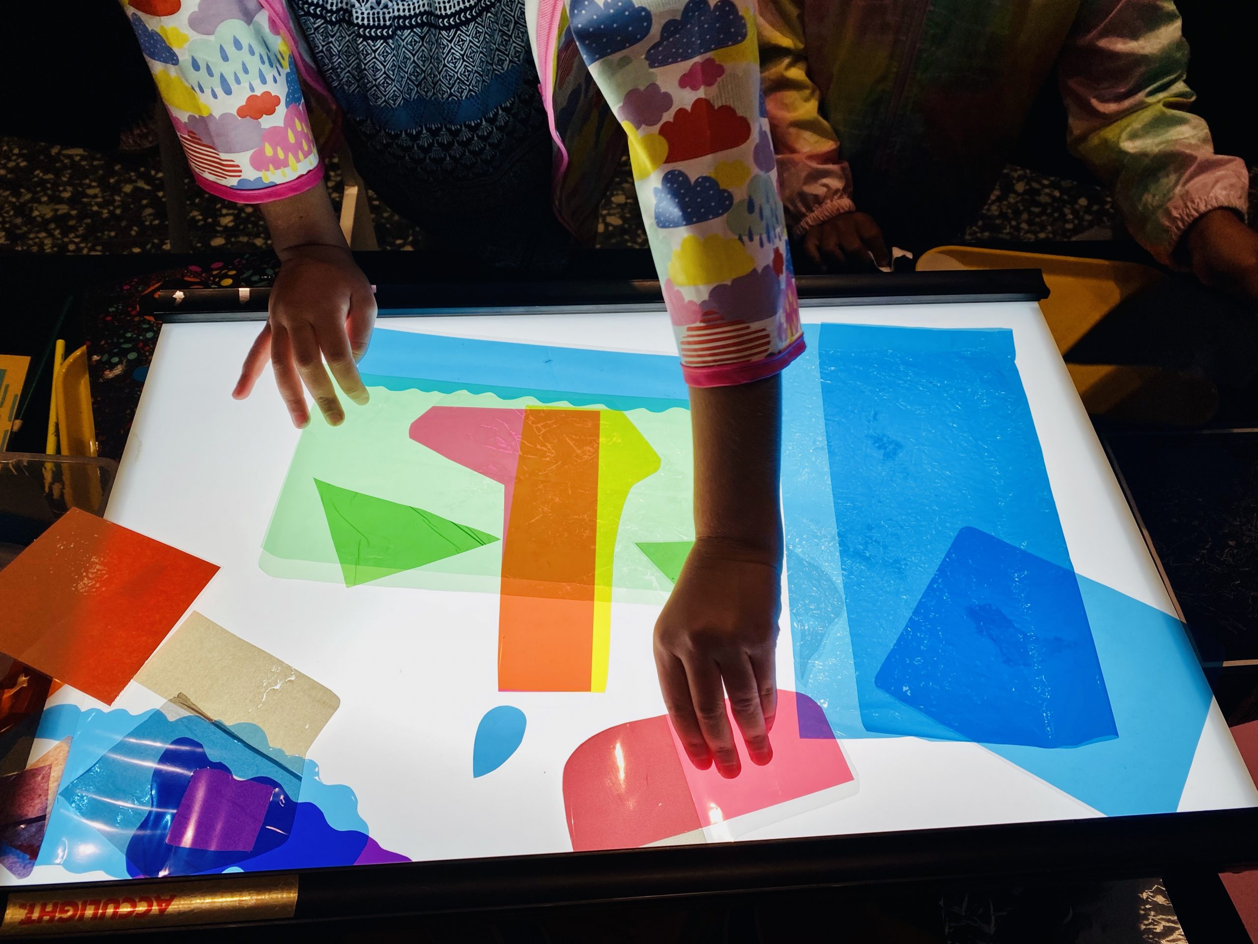 A child's hand places a small pink piece of paper on top of a light box.