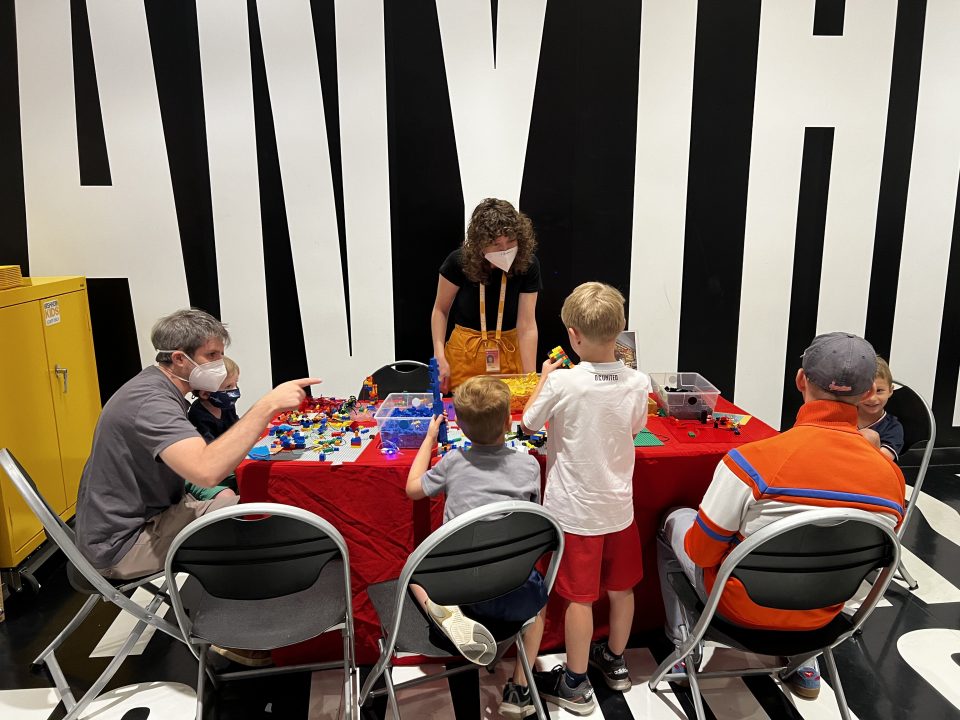 An educator speaks with children and their families at a lego craft table in the lower level, surrounded by Barbara Kruger's Belief+Doubt