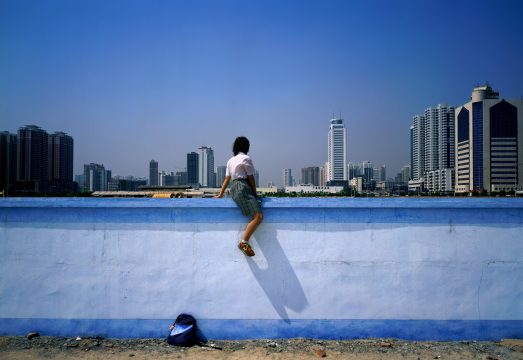 A young woman sits atop a wall against a city skyscape