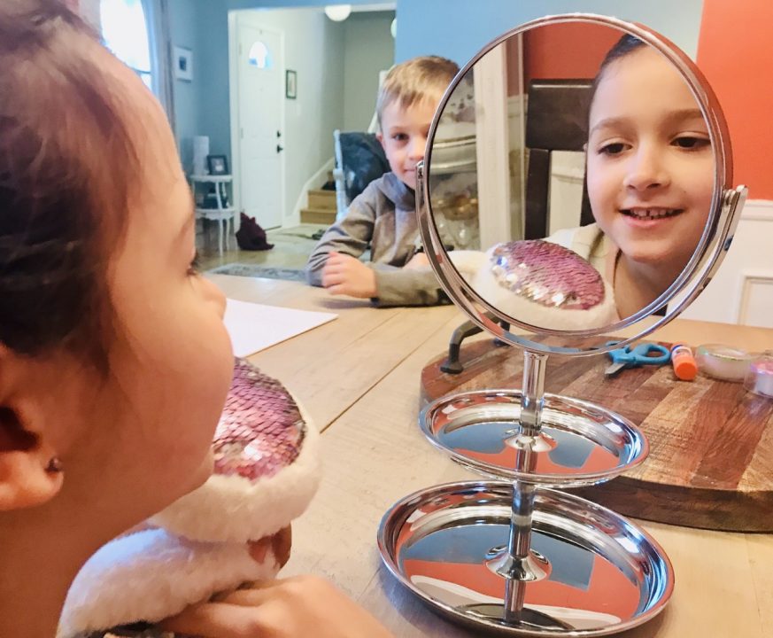 A child in the foreground facing away looking in a mirror in the background is a mirror with her reflection and another child looking on.