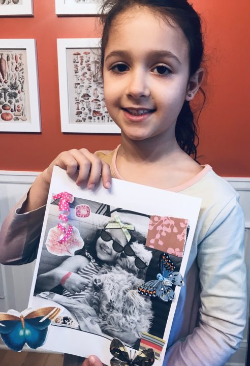 A young girl holding up her self portrait in front of her chest smiling. She is standing in front of an orange/red wall with colorful prints hung up