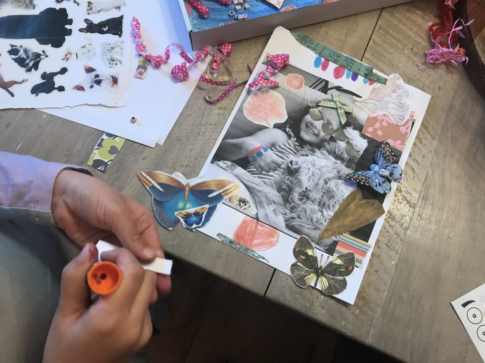 A child’s hands in the foreground applying glue to a collage piece. In the background is her portrait with several colorful additions and butterflies