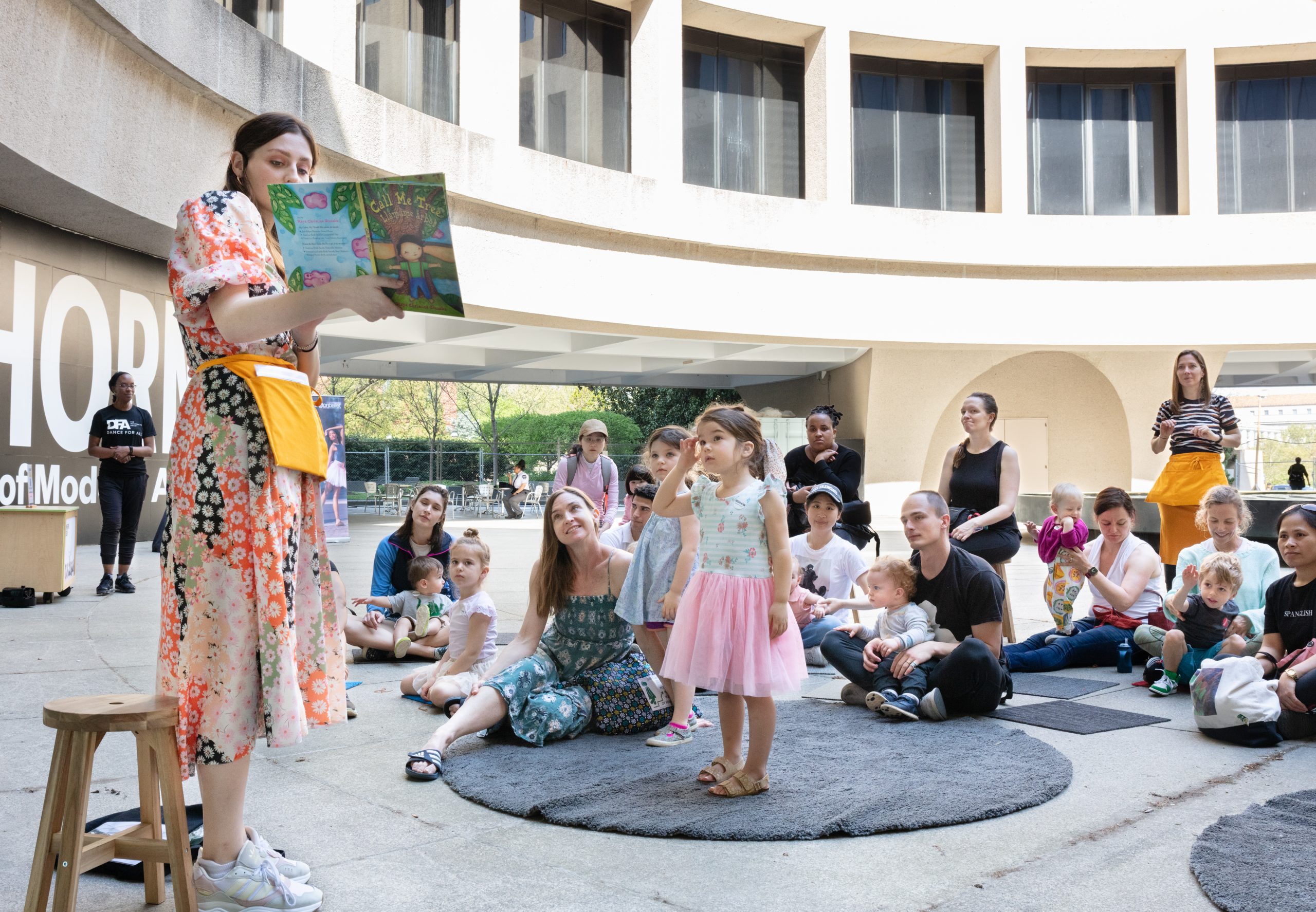 An educator stands in the Hirshhorn's plaza holding up the children's book Call Me Tree and reading it to parents, caregivers and children assembled on round and square grey mats.