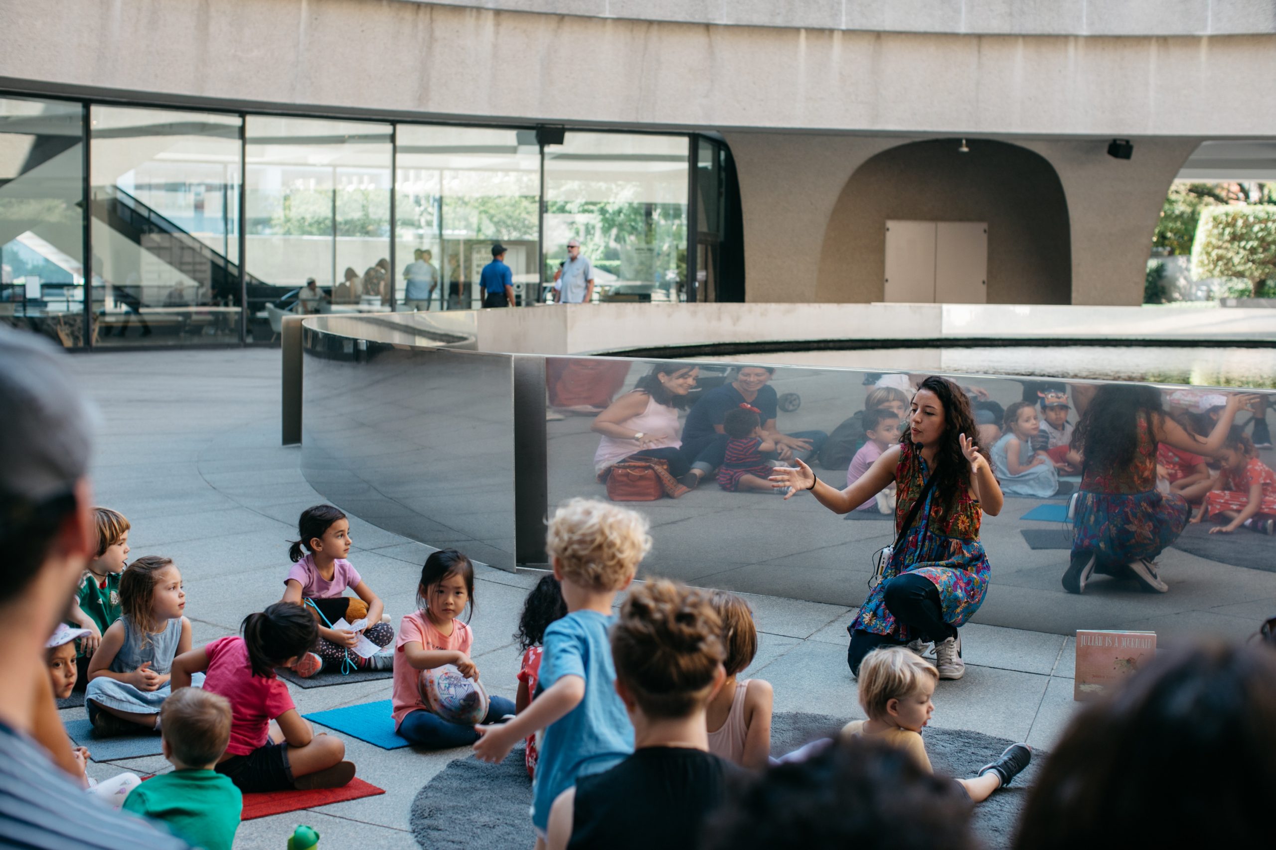 An educator kneels down in front of the fountain on the Hirshhorn Museum's plaza, animatedly telling a story to a group of youngsters and their adult caretakers.