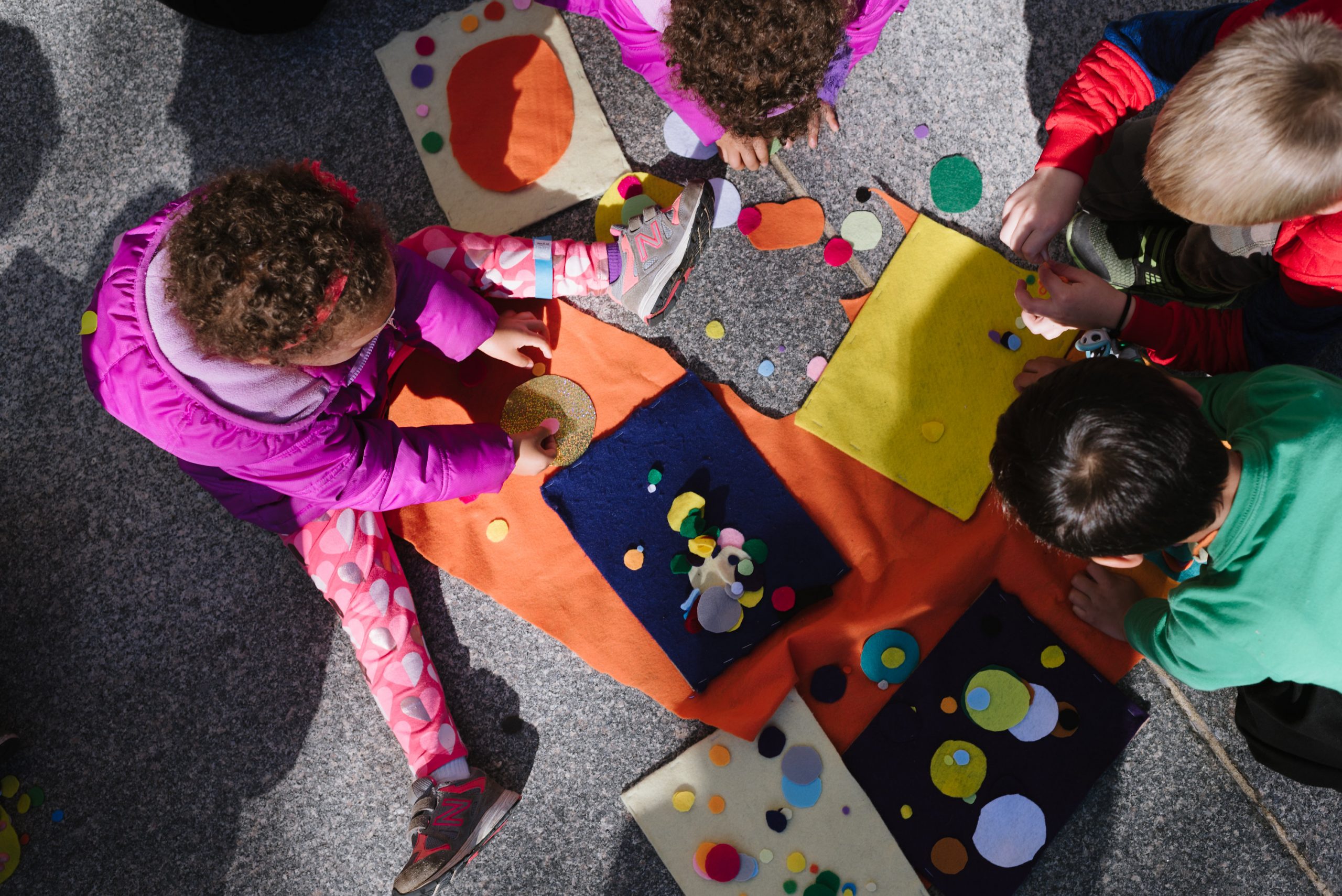 A birds eye view of four children, gathered together in a circle creating felt artwork by attaching small organic shaped felt pieces to larger, square bases.