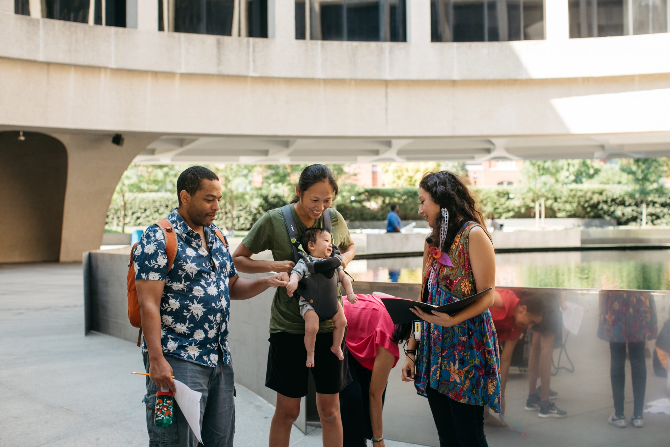 A group of three adults are gathered around the shiny metal sheet surrounding the museum's fountain the the plaza of the Hirshhorn.