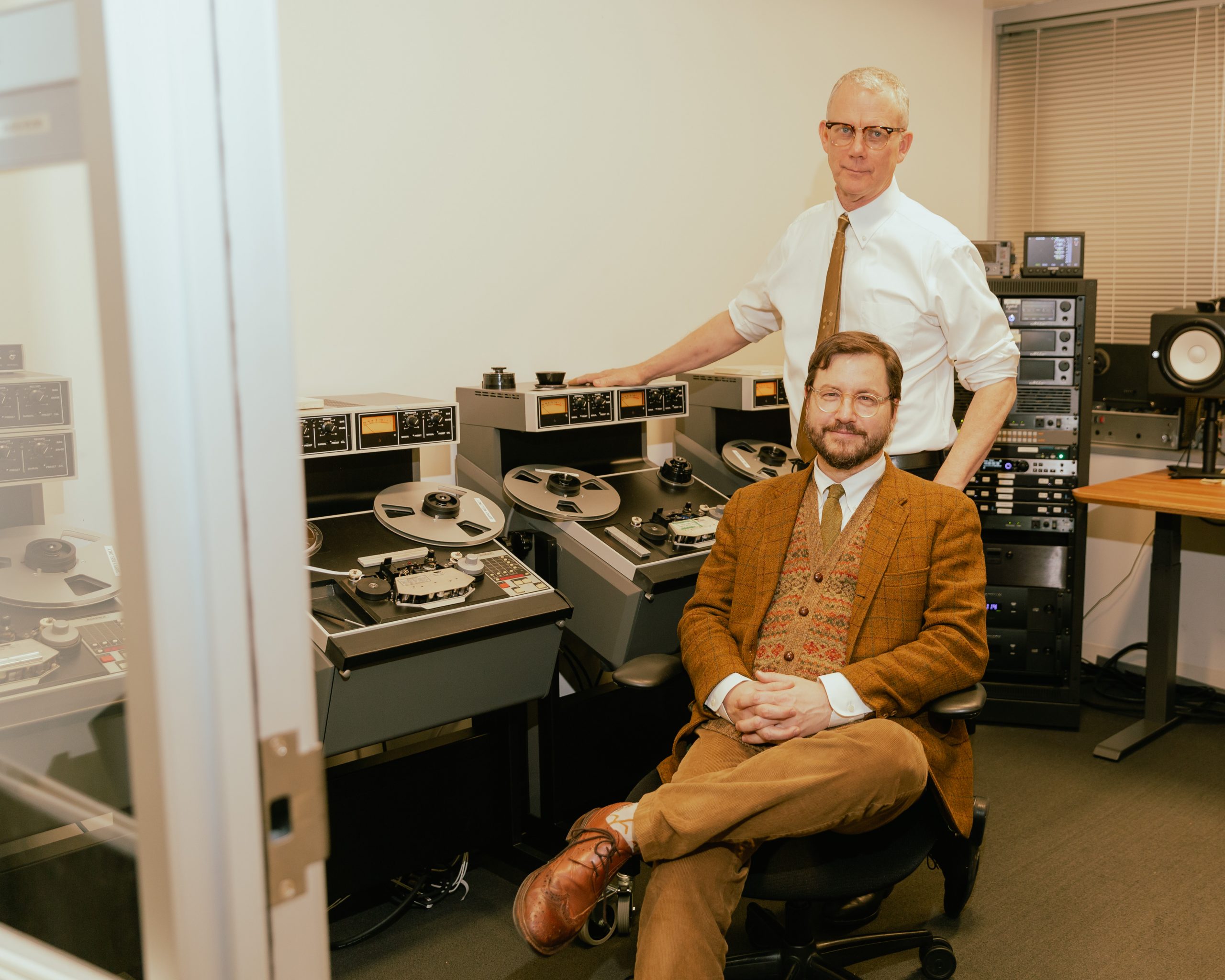 A man in brown sitting in a chair and man in white standing behind him next to a recording a machine.