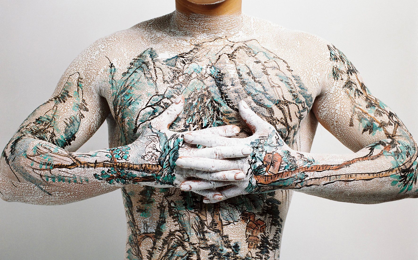 an image of a man with a painting of a mountain on his chest. his arms are bent and his hands interlock in the middle of his chest.