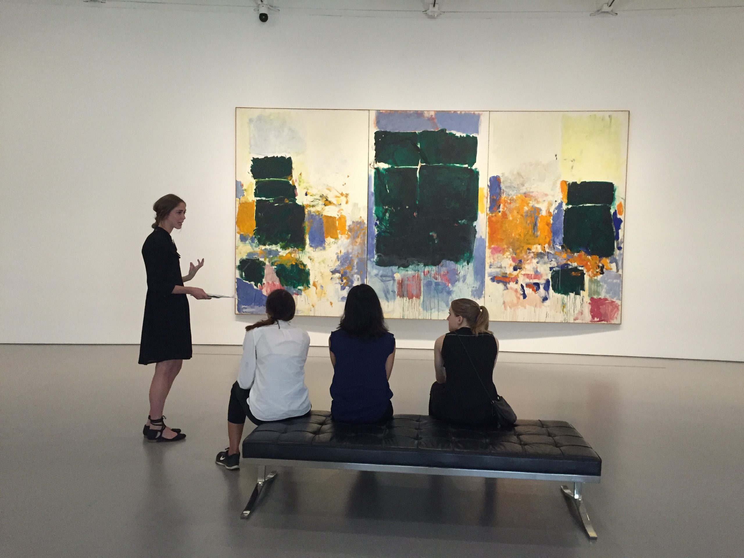 a woman in a black dress is speaking with three women who are seated on a bench in front of a colorful triptych painting by Joan Mitchell