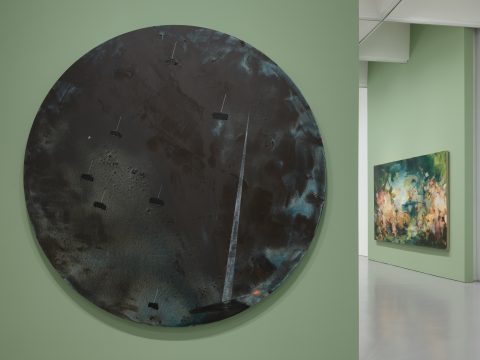 Two abstract paintings display in a gallery. The one of the left takes up most of the space, it is a large black, blue and grey circle. On the right is another abstract painting, rectangular in shape and features abstract strokes of blue, green, peach and brown colors.