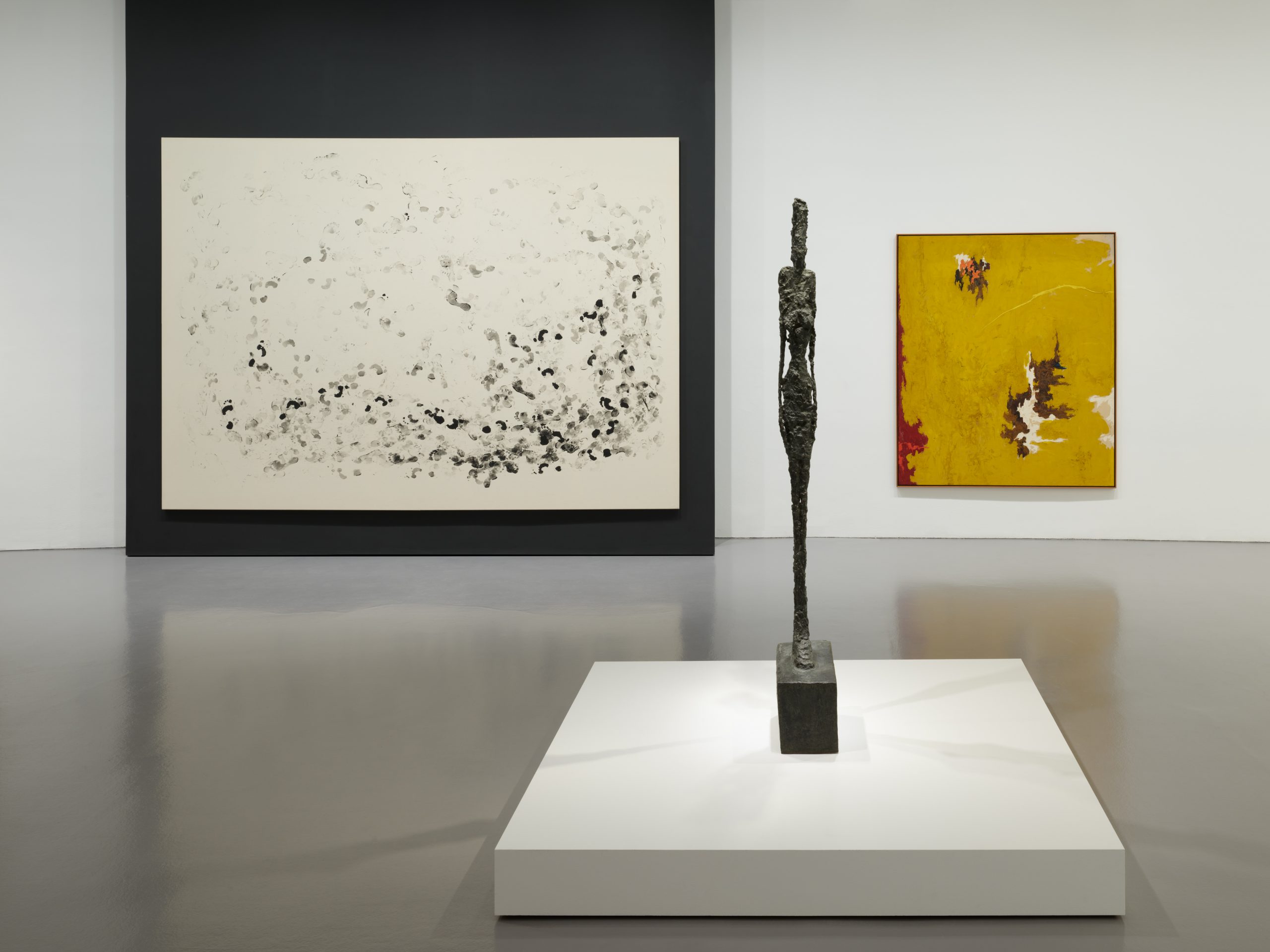 Interior view of an art gallery showcasing two abstract paintings on the wall and a sculpture of a man in between the works.