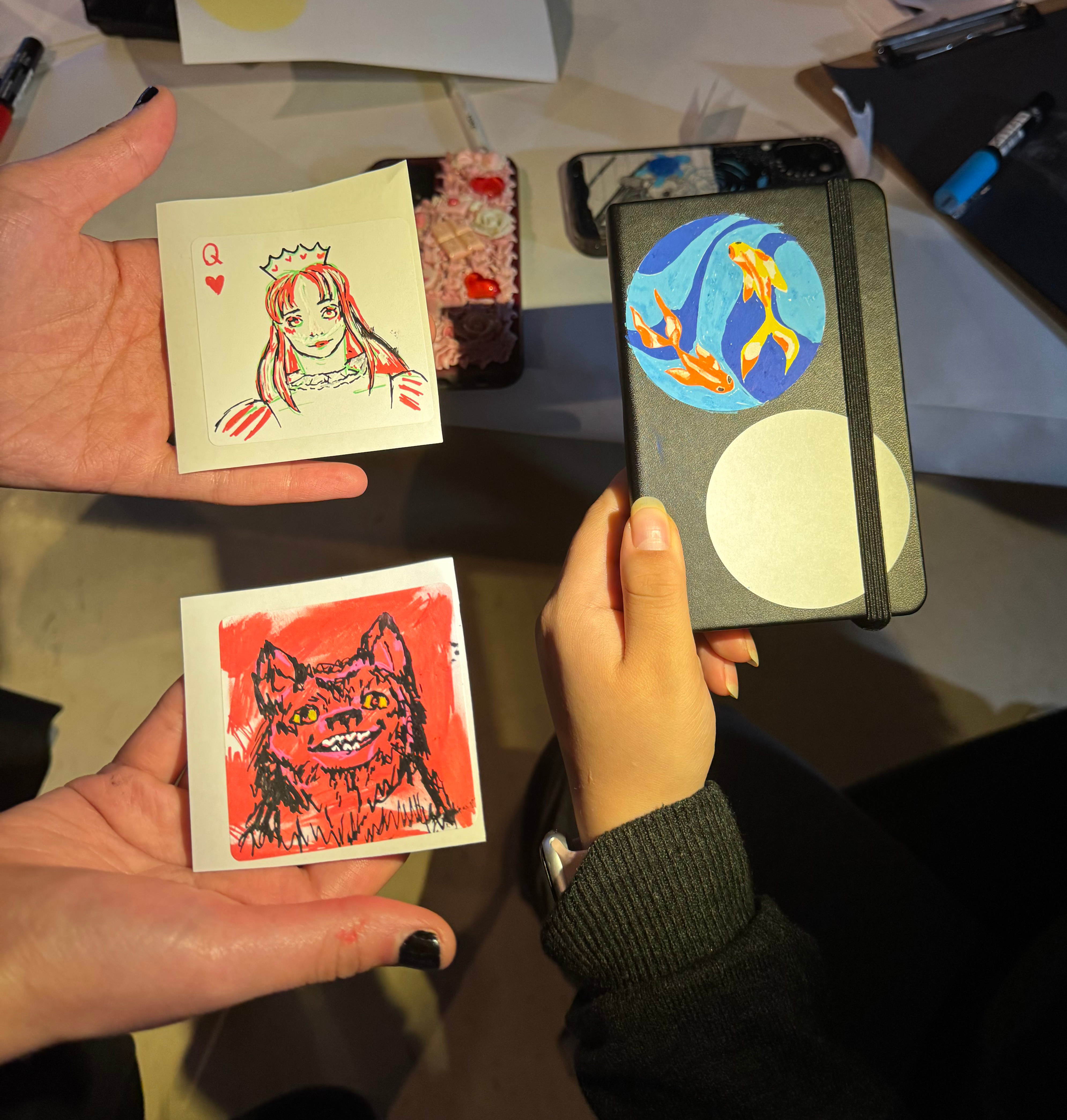 a group of teens holding individual drawings. from the top left, clockwise: an image of a playing card displaying a Queen, a notebook with two koi fish on the cover and an image of a red wolf.