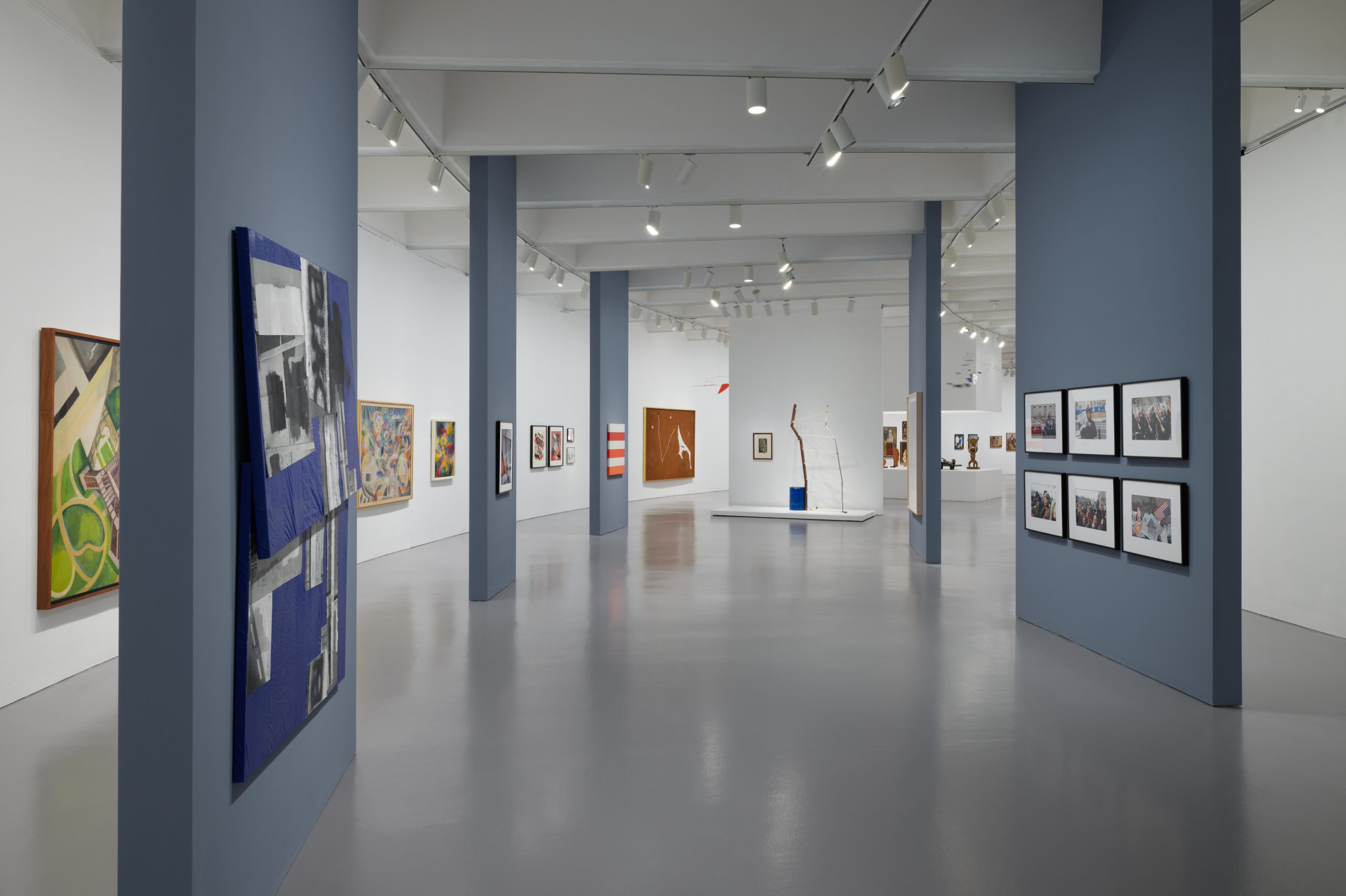 a hallway in the gallery, featuring five blue blade walls with contemporary art on them. The walls surrounding the blade walls feature colorful modern art.