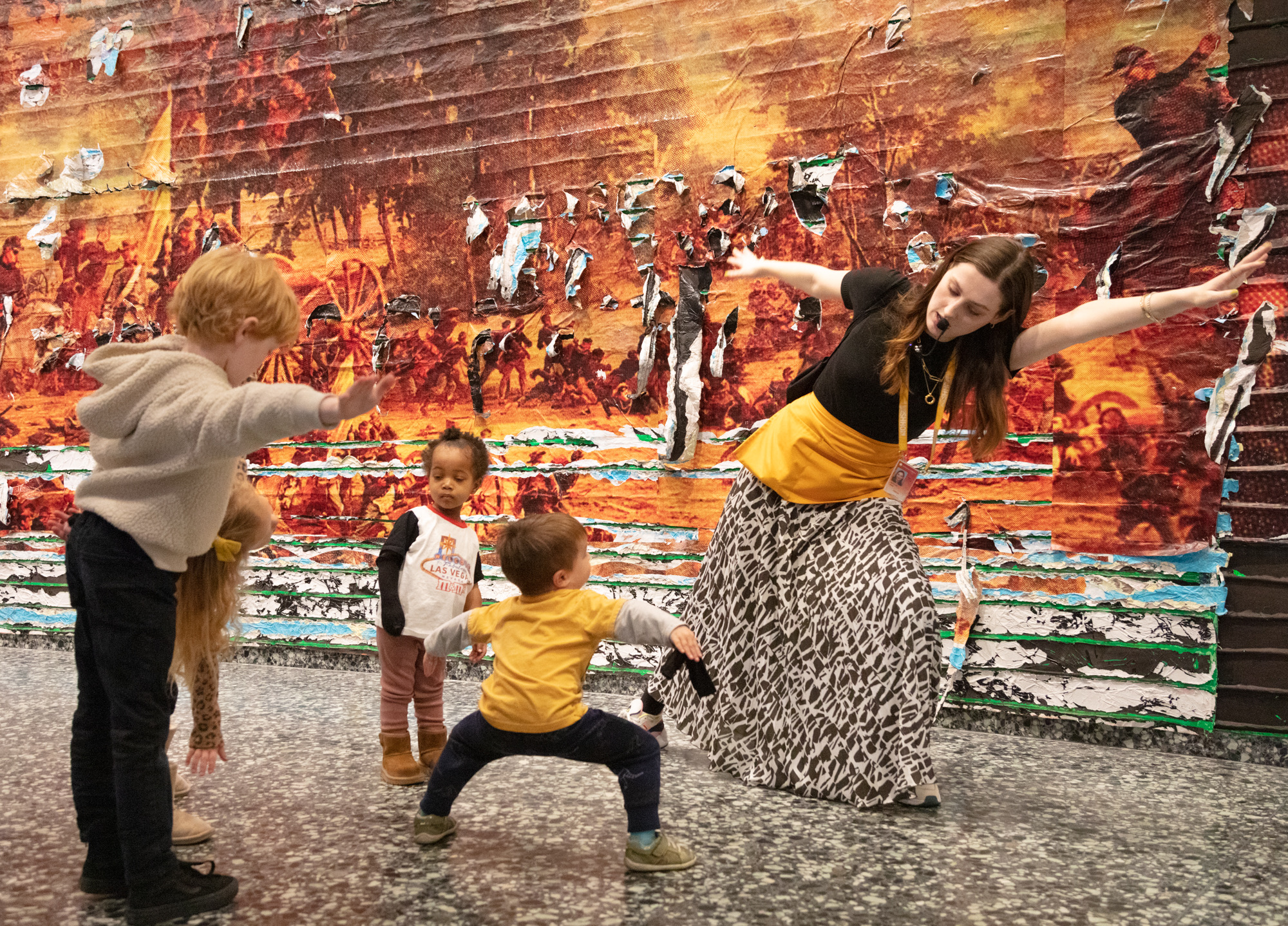 a young woman with a microphone is stretching her arms out with four young children mimicking her moves in an art gallery