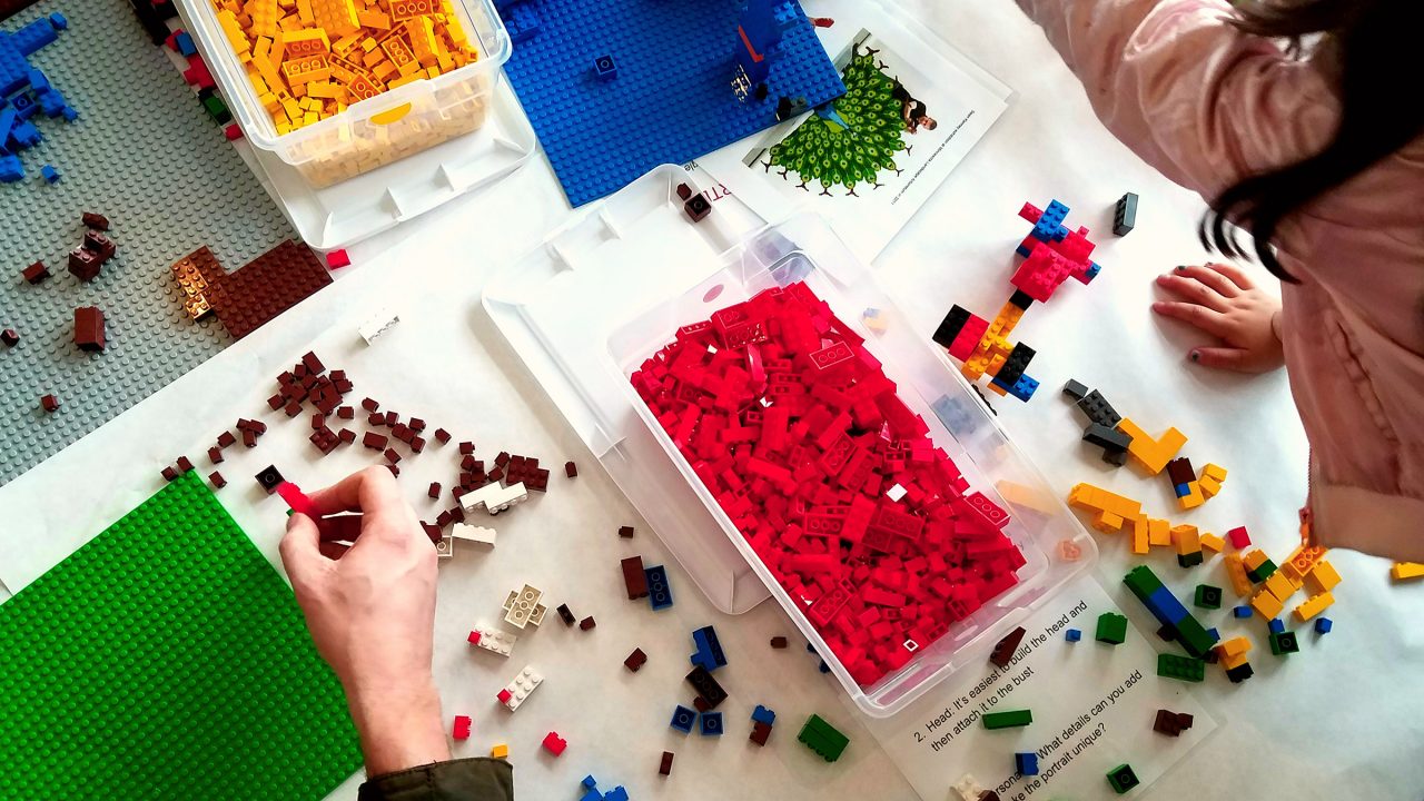 several colorful boxes of legos are on a white table laid out. Children and adult hands are assembling the pieces.