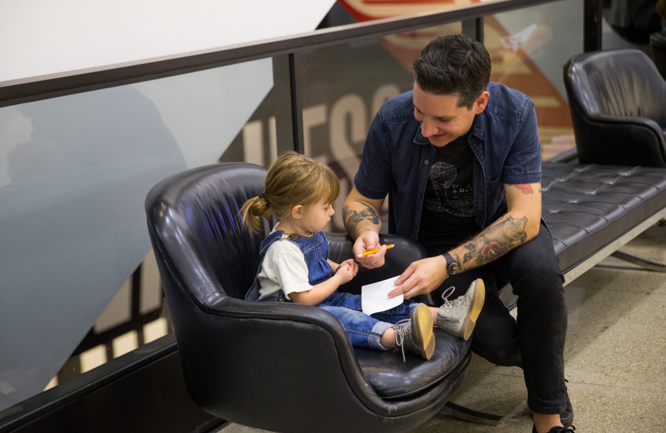 A small child with pigtails and overalls sits in a black leather chair, holding a pencil in her chubby hand. A man sitting next to her is smiling and holding a piece of paper over her body.