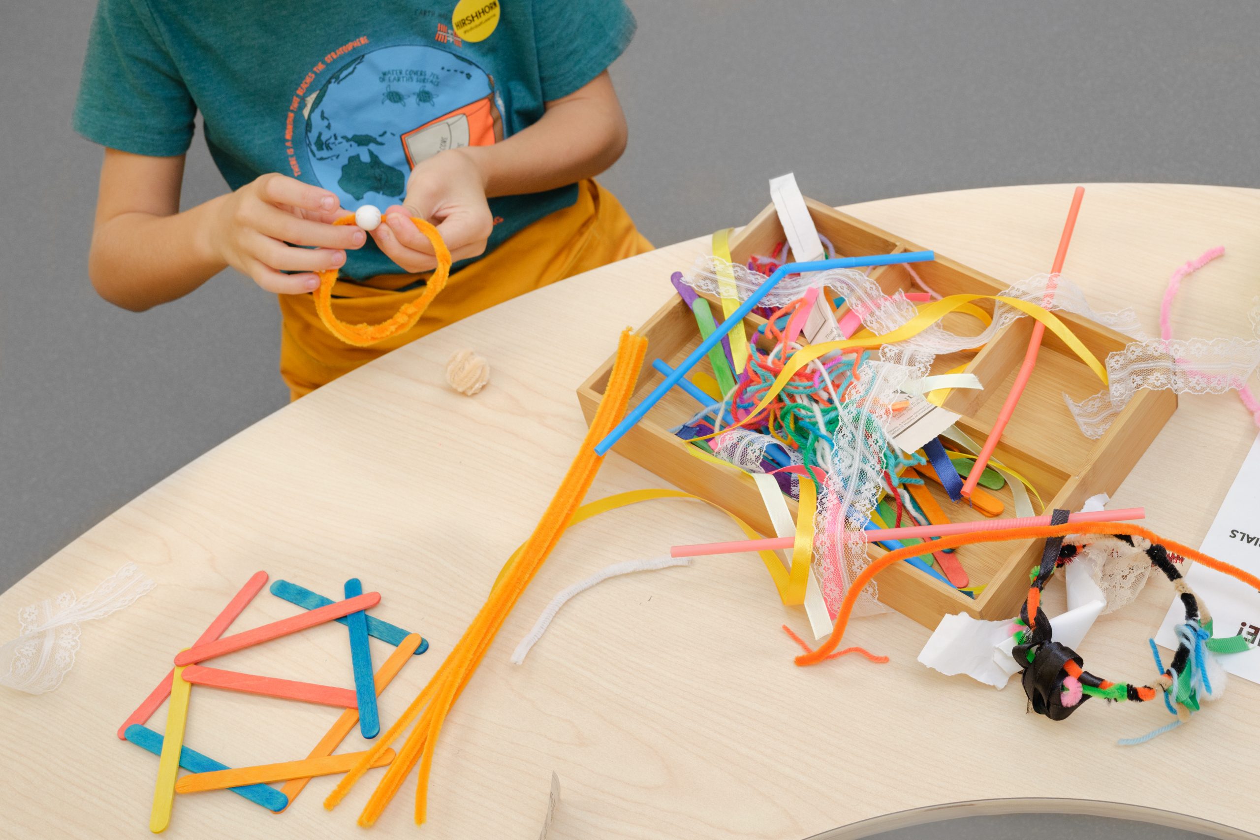 A young child sits at a curved table. A tray on the table is filled with colorful art supplies including string, paper strips, and pipe cleaners.