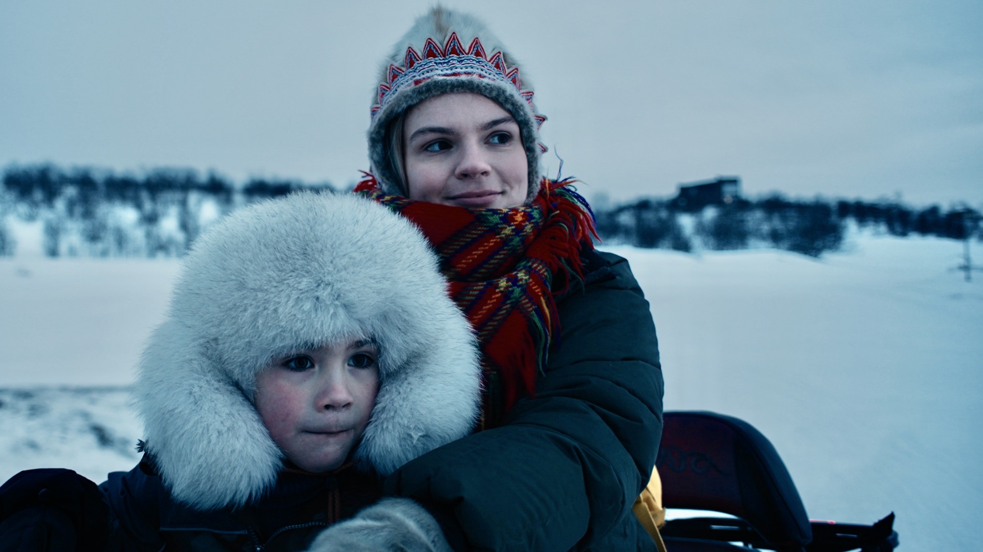 two young women on a sled in the middle of a snowy location are dressed in winter gear and look away from the camera