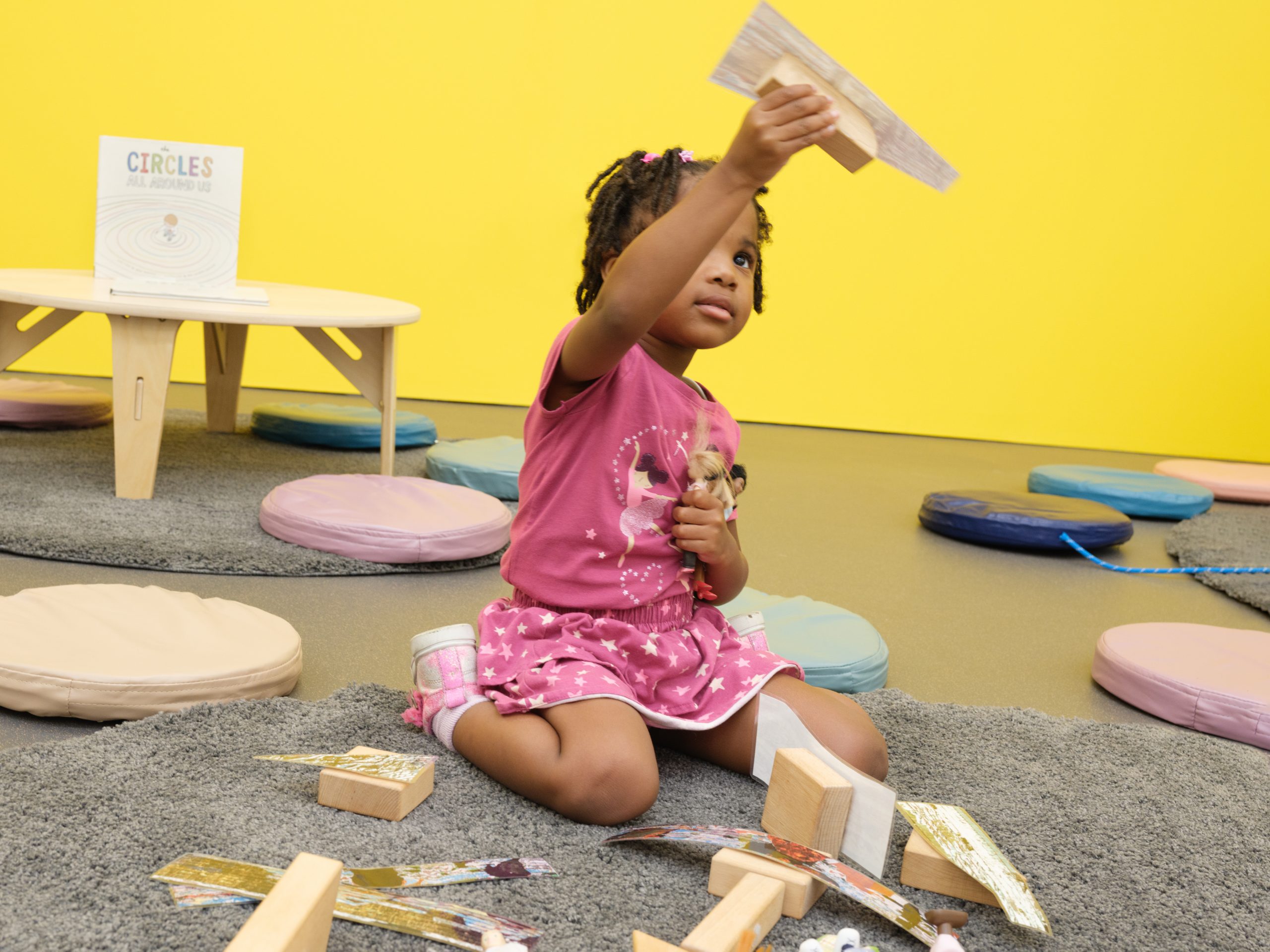A child sits on her knees a gray floor rug surrounded by blocks and colorful circle cushions. The child holds a paper airplane in the air.
