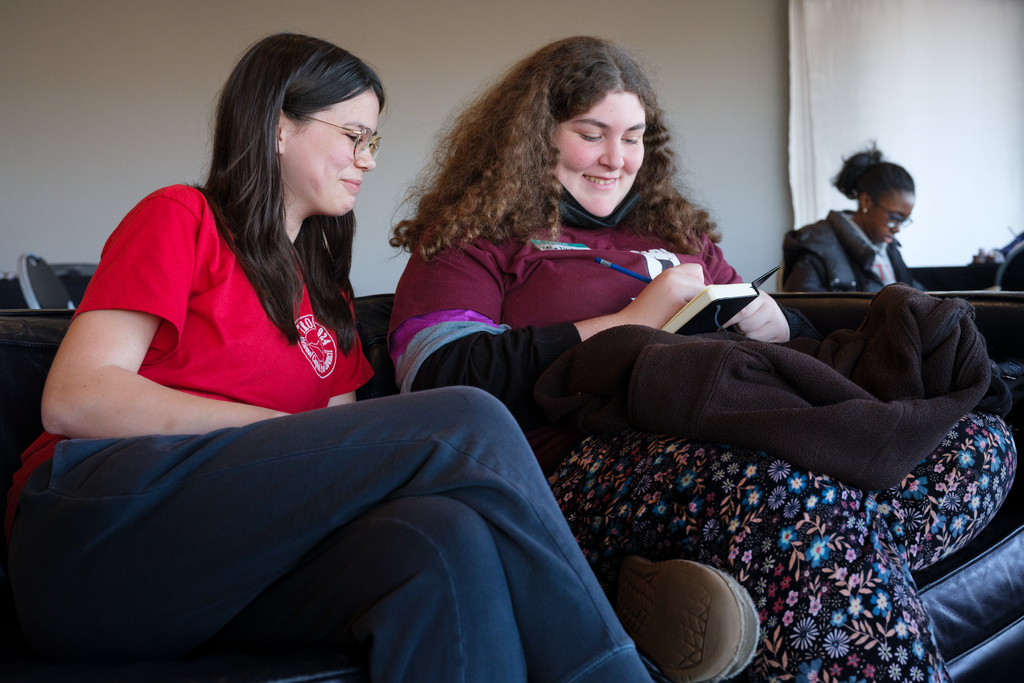 Two young women seated on a couch, focused on a book and smiling.