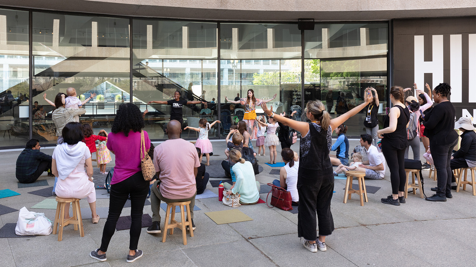 Individuals are seated and standing during a children's activity in front of a building displaying a Hirshhorn sign enjoying the outdoors together