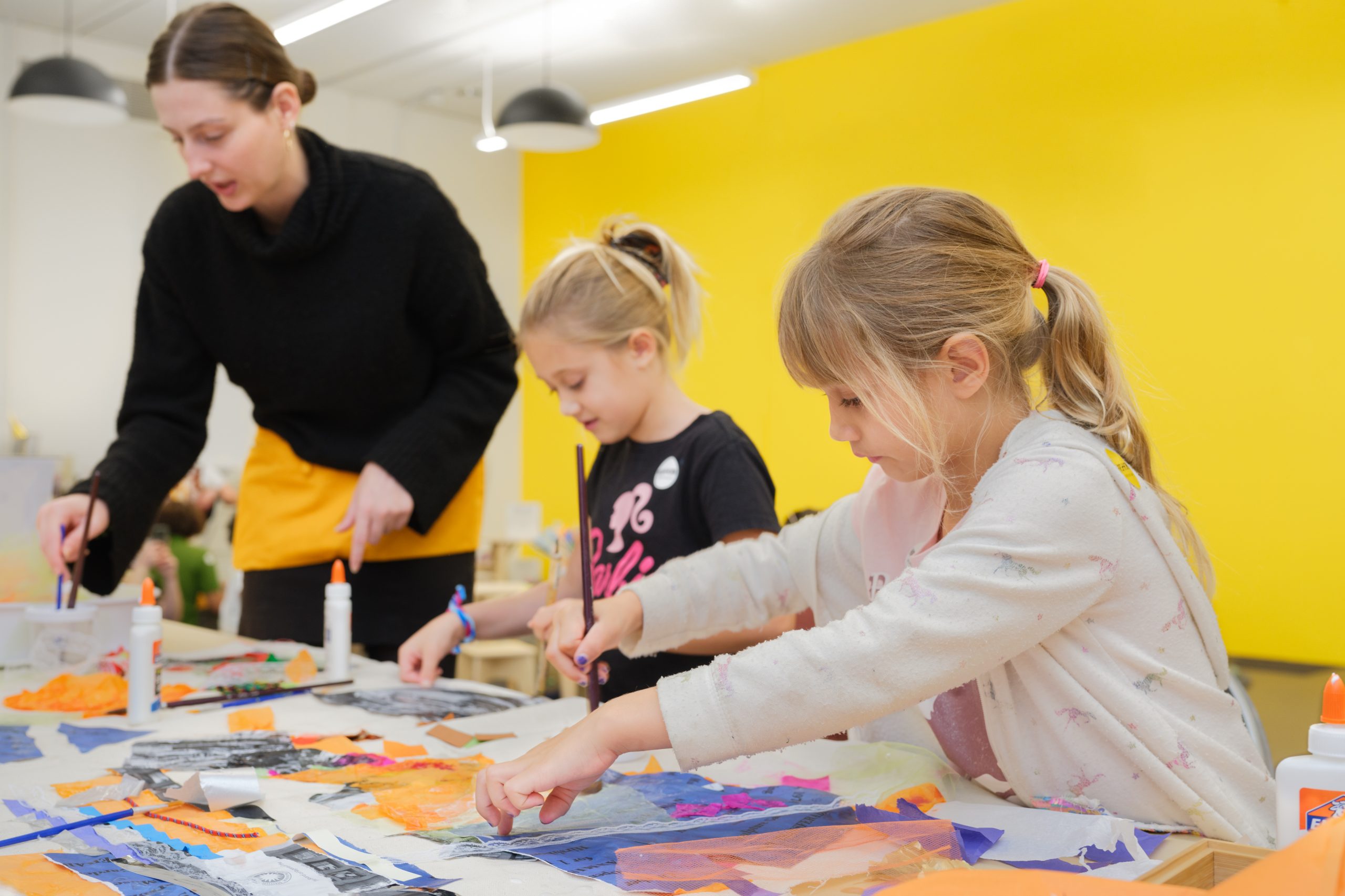 Two children and a woman stand at a table covered in art materials, including glue and scraps of blue, orange, and black paper. The child at left and the woman use paintbrushes to glue down paper scraps, while the child at right arranges the scraps with her hands.