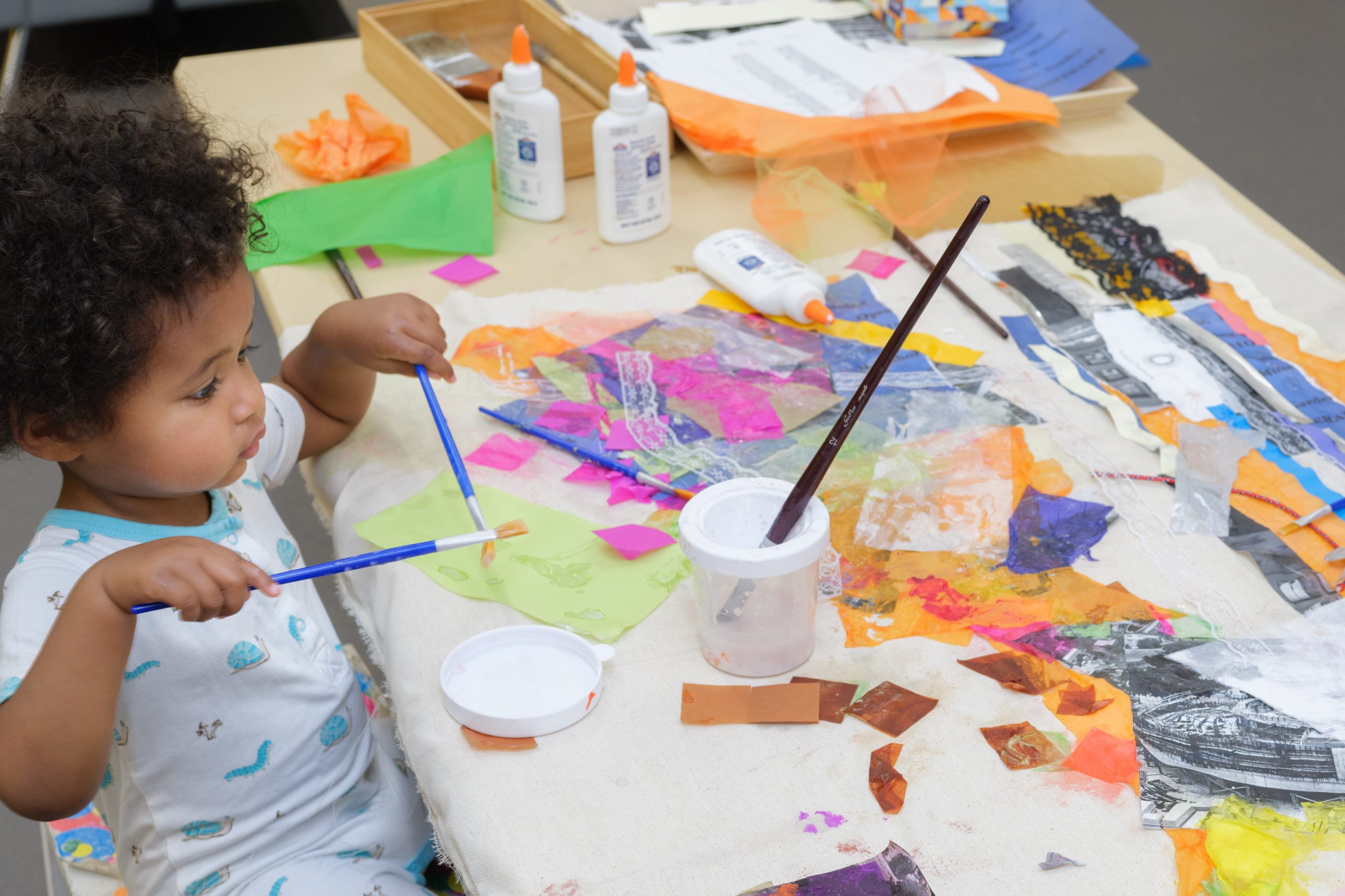 A child holding two paint brushes applies glue to an artwork, already covered with scraps of colorful tissue paper and black and white photographs, on the table in front of him