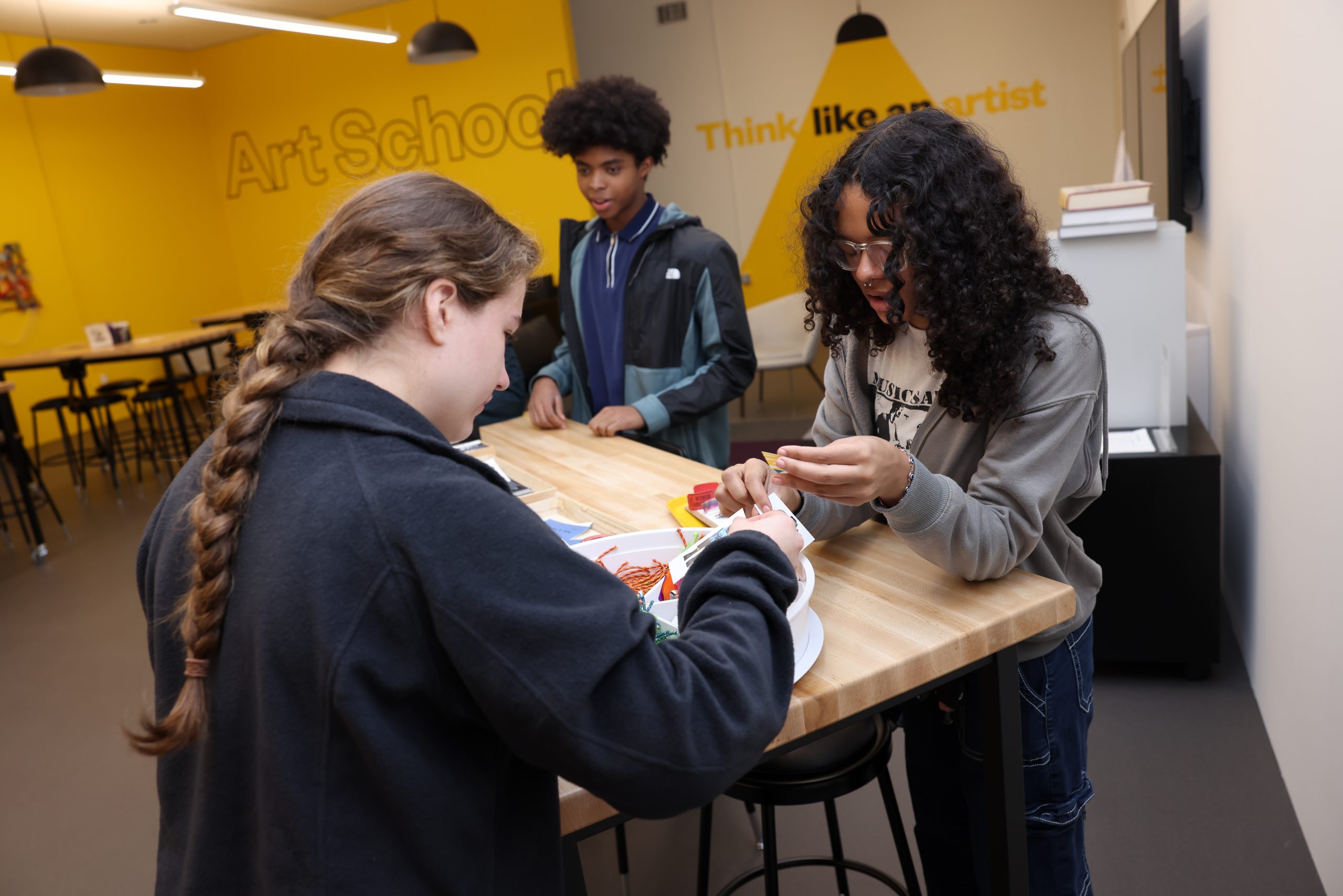 In a bright yellow room, three teens stand at a work table on which sit tape, rope, pieces of newspaper, and glue in a white container