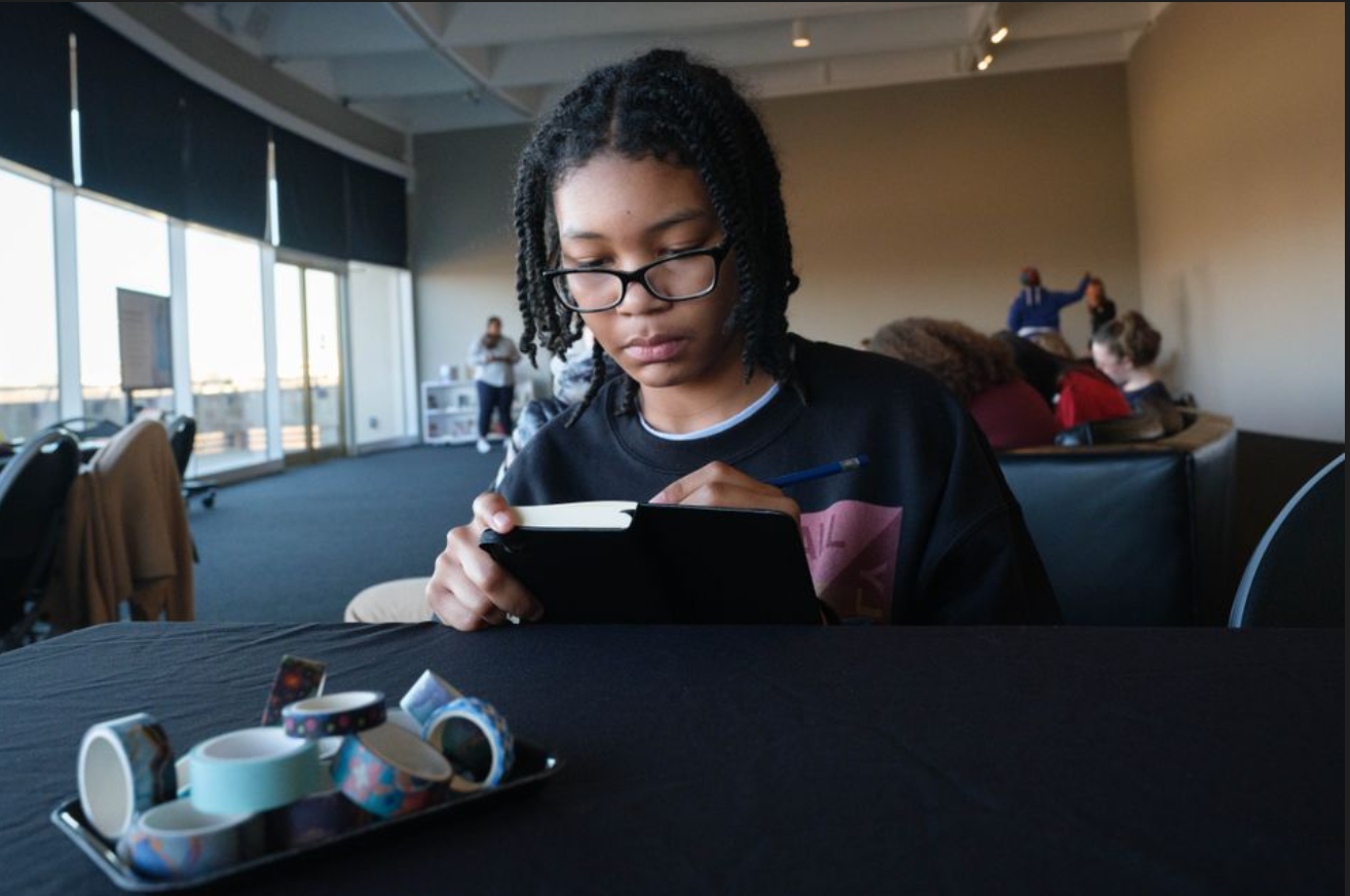 In a window-walled room, a teen sits at a black table, writing in a small black notebook with a pencil. On the table sits a tray of tape.
