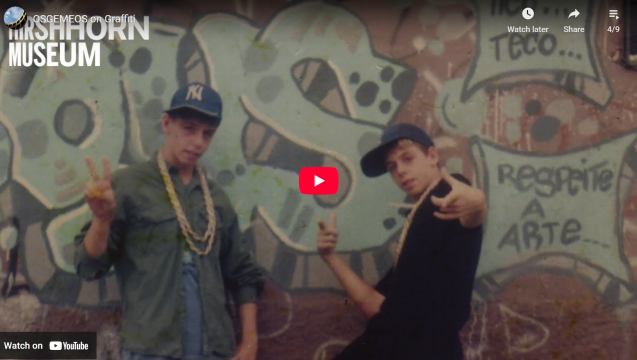 two young boys pose in front of light blue, spray painted graffiti. The boys are wearing long gold chains and blue baseball caps. 