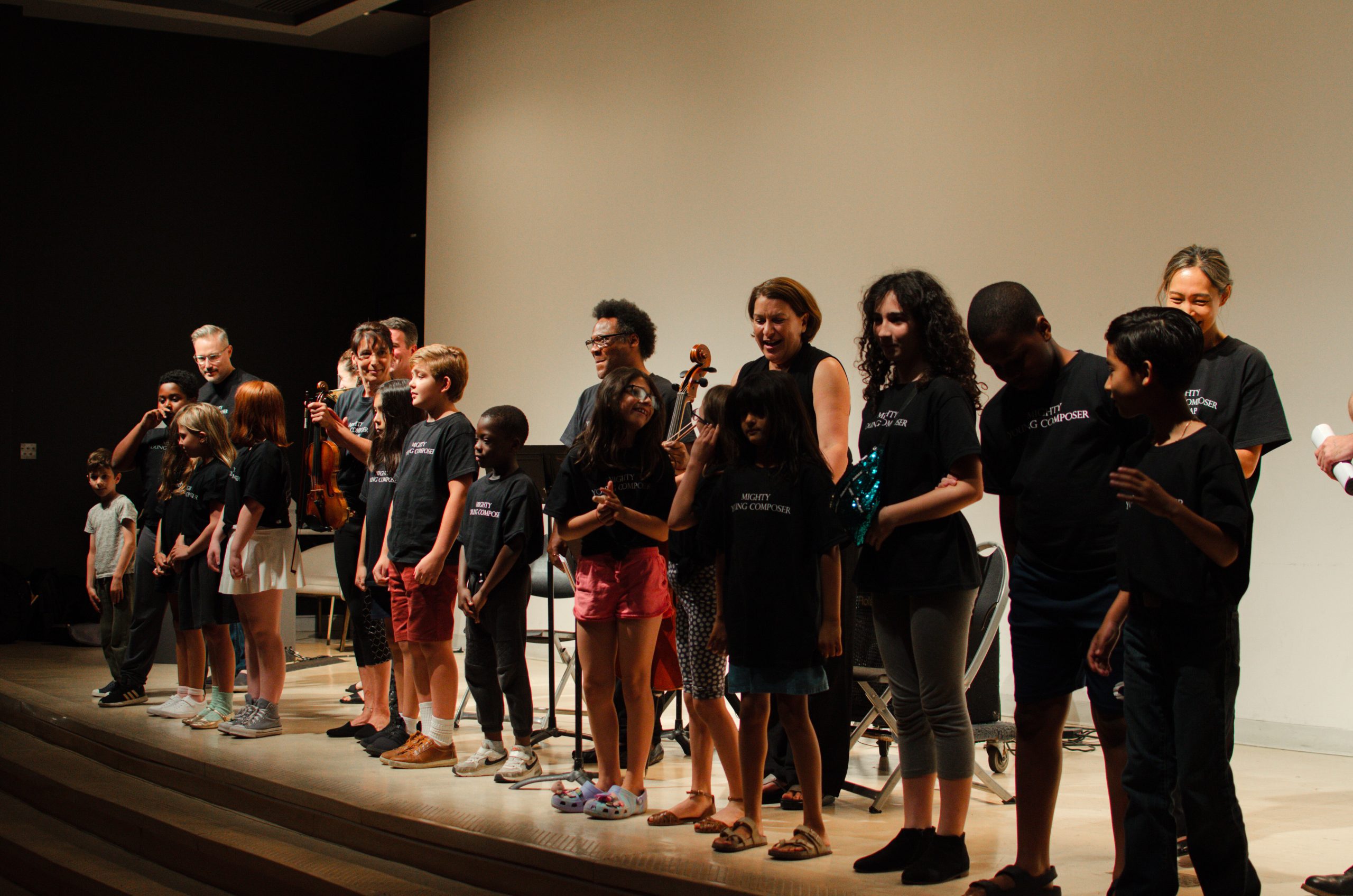 A group of children and adults stand on a stage in black "Mighty Young Composer" shirts, smiling with musical instruments, conveying a joyful, celebratory mood.