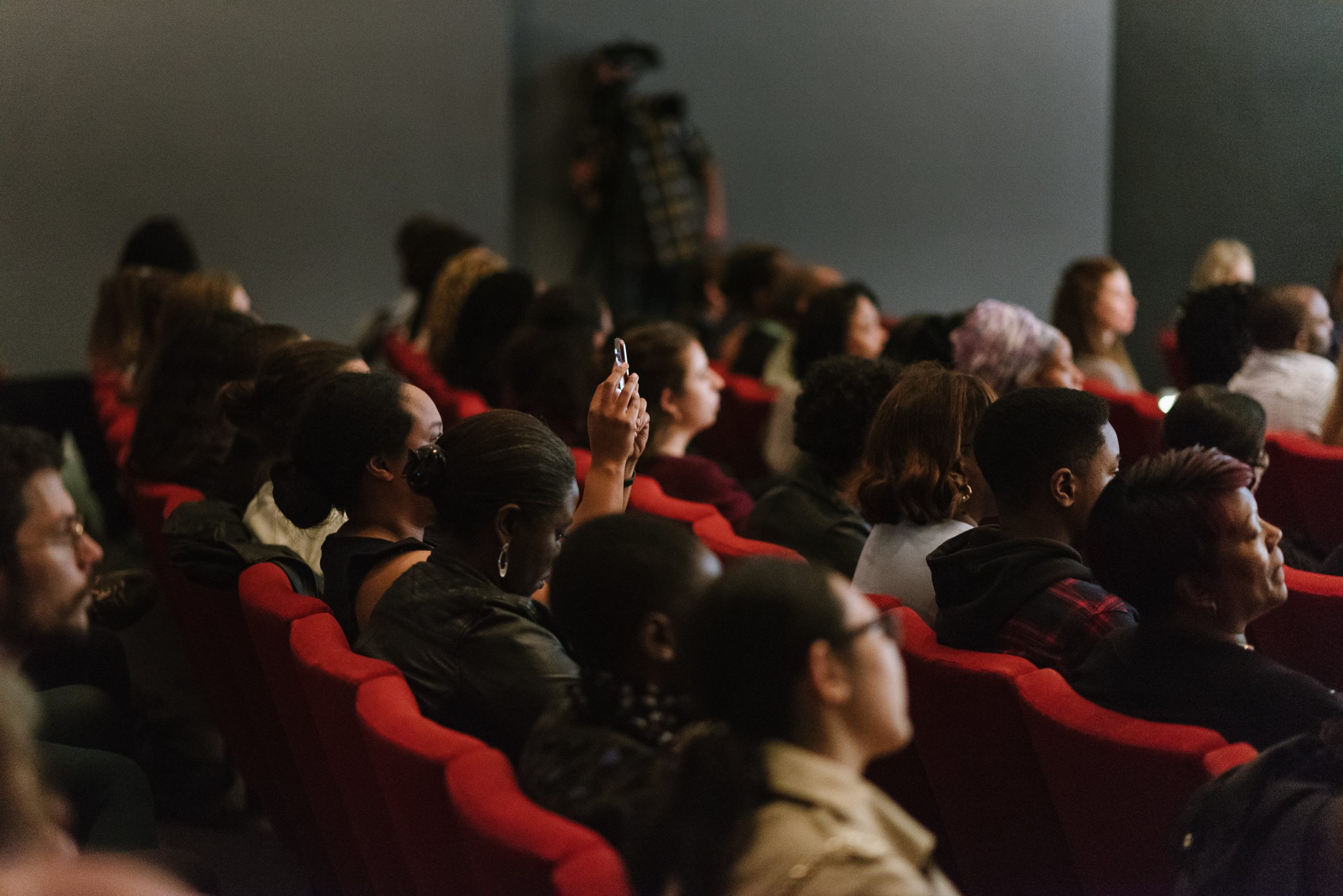 people are sitting in a dark auditorium with red seats. You can see the back of their heads. One person is holding a phone up in the air.